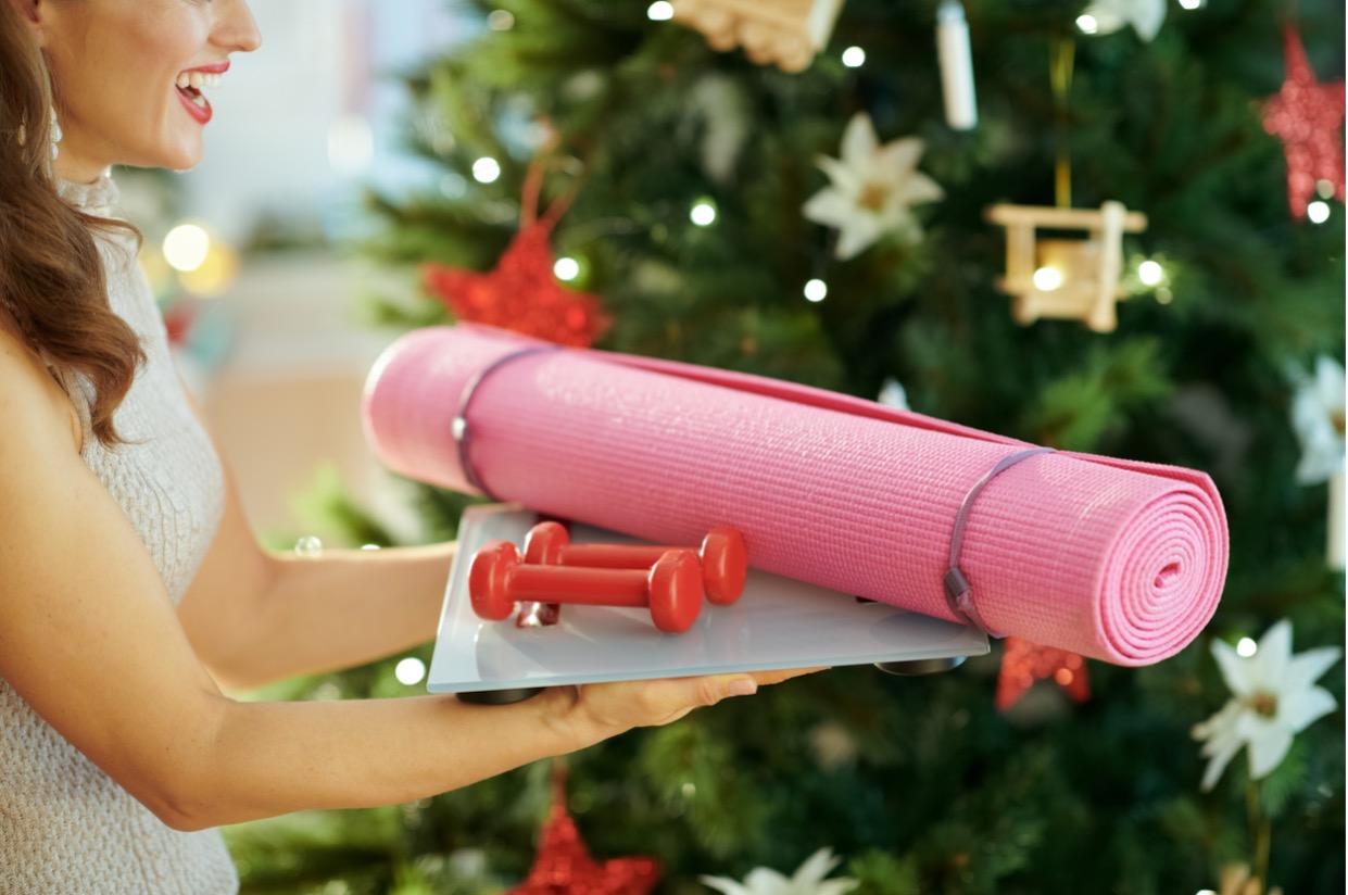 cheerful lady holding exercise mat and small weights infront of christmas tree