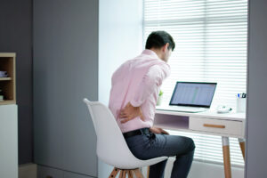 Back Pain from Desk Job? Here’s How to Fix It, A person sitting at their desk with their hand on their lower back in pain