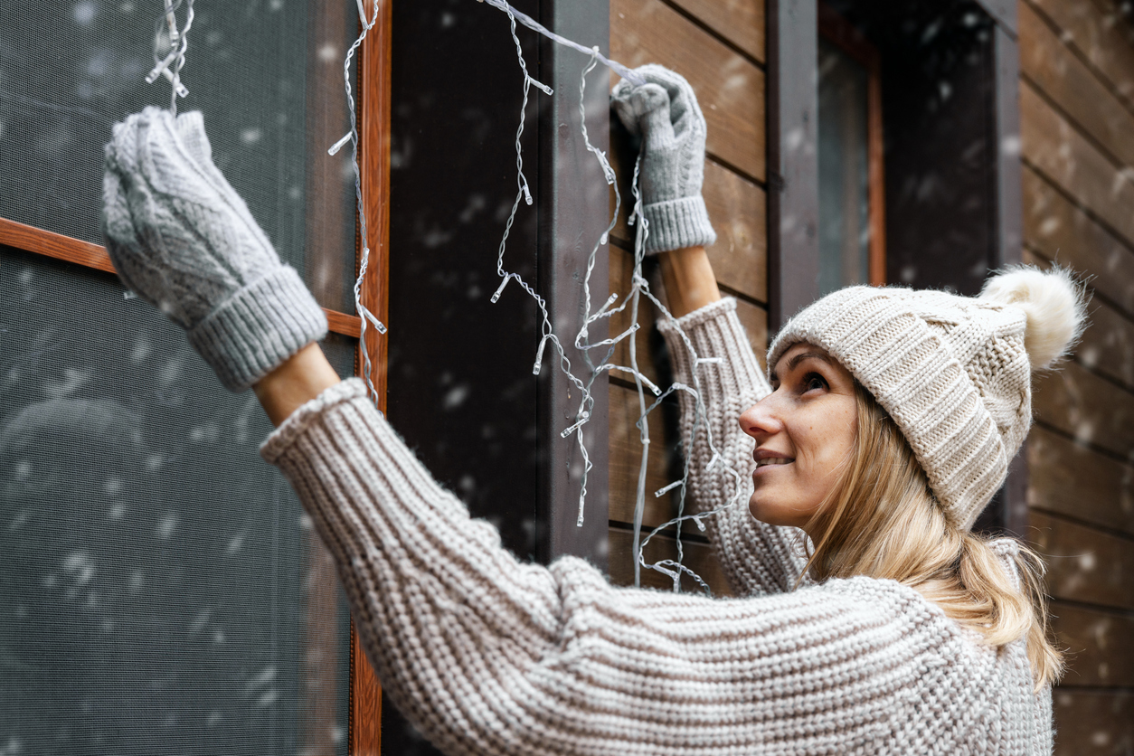 holiday decorations. woman install electric christmas string lights on house exterior facade, holiday survival tips for a healthy spine