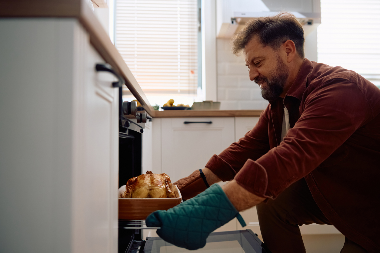 Smiling man taking roast turkey out of the oven while preparing food for Thanksgiving. Copy space, holiday survival tips for a healthy spine