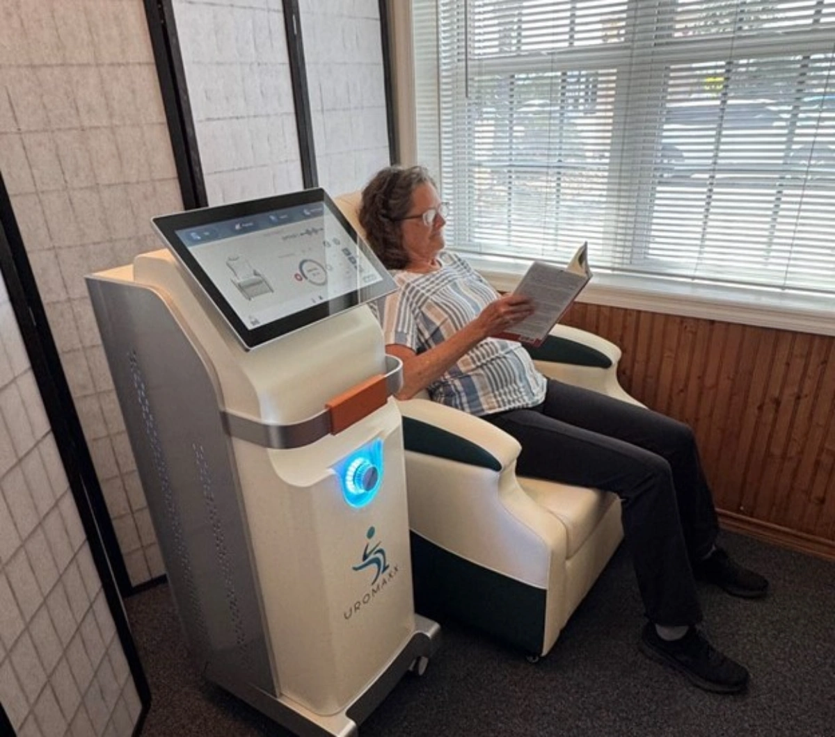 Woman reading book while sitting in a UROMANXX treatment chair next to a medical device.